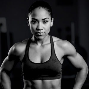 This image shows a black and white portrait of a fit African American woman wearing a sports bra in a gym setting. Her expression is focused and determined, highlighting her strong, toned physique. The lighting accentuates her facial features and muscles, creating a dramatic and powerful visual.